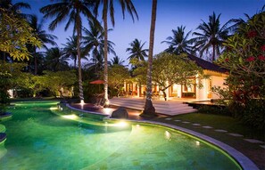 Zona de piscina con borde curvo, palmeras y un edificio con tejado de baldosas.