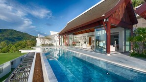 A modern house with a large swimming pool, a covered outdoor area, and a view of a green hill.
