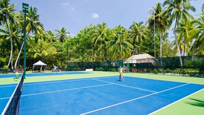 Un court de tennis où s’affrontent deux joueurs, entouré de palmiers et surmonté d’une structure à toit de chaume.