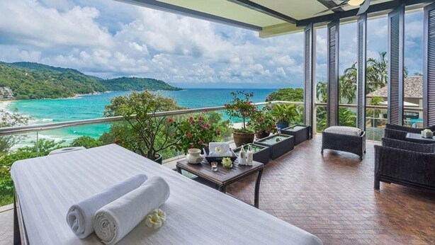 Ein Balkon mit Bett, Tisch und Blumentöpfen, der auf eine Küstenlandschaft mit Blick auf das Meer und die Hügel blickt.
