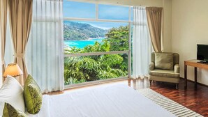 Ein Hotelzimmer mit großem Fenster und Blick auf eine Küstenlandschaft, einem Bett, einem Sessel und einem Fernseher auf einem Holztisch.