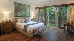 Een slaapkamer met een groot bed, houten nachtkastjes, een bankje en uitzicht op een tropische tuin door een glazen deur.