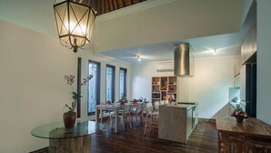 Een ruime woonkamer met een eettafel, stoelen, een salontafel met glazen blad, en een keuken met fornuis en kasten.