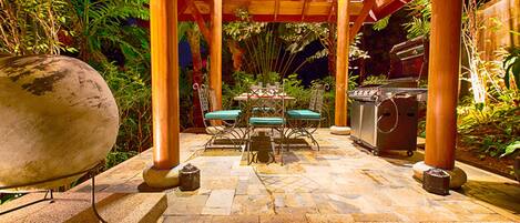An outdoor patio with a stone floor, a round grill, a table with chairs, and a wooden pergola structure.