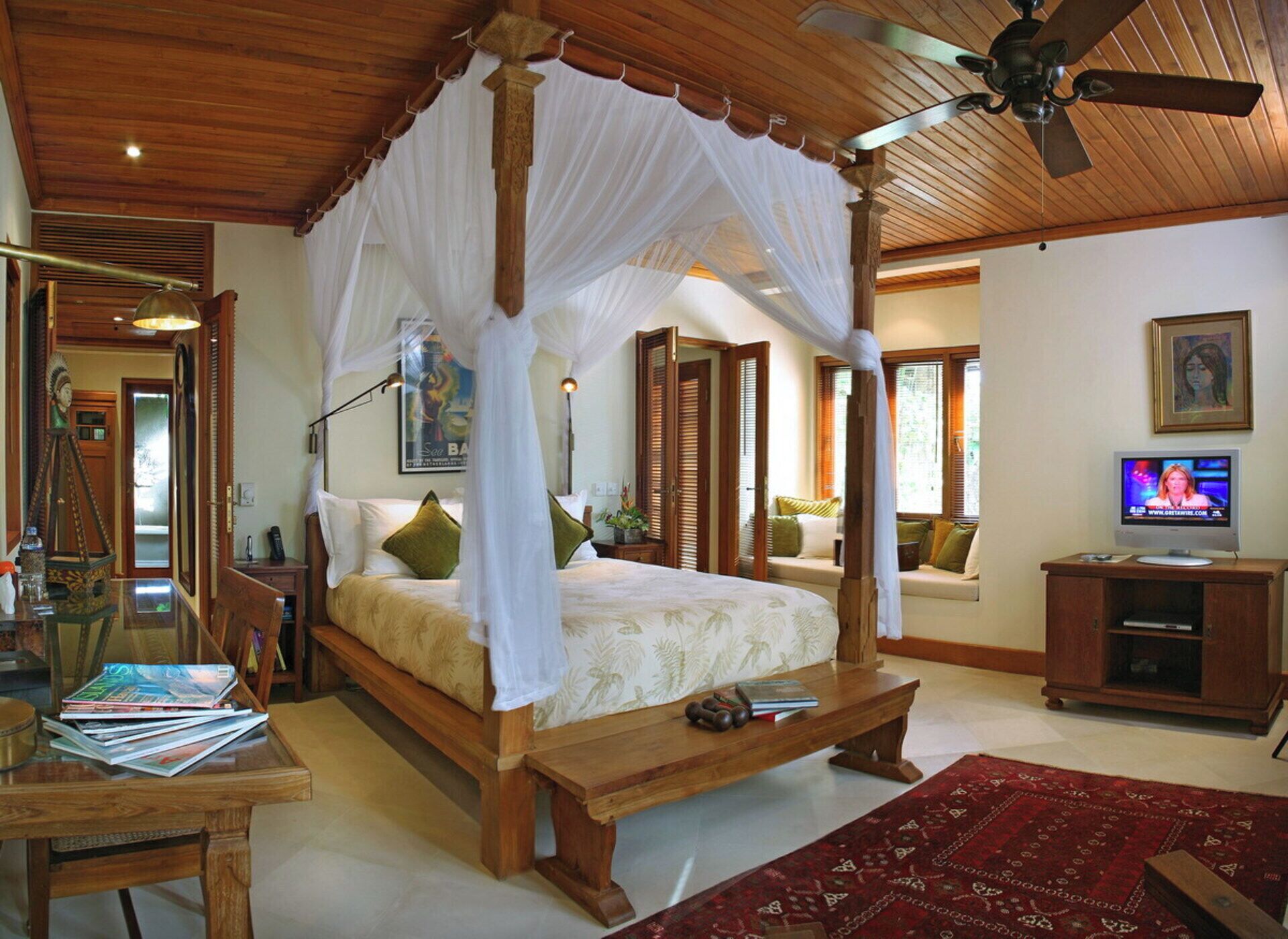 A four-poster canopy bed with a wooden frame and white bedding, a wooden desk with magazines, a television on a wooden cabinet, and a red patterned rug on the floor.