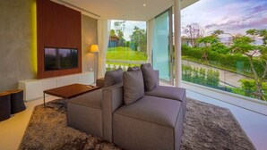 Una sala de estar moderna con un sofá gris, un televisor de pantalla plana montado en un panel de madera, una pequeña mesa de centro de madera y un amplio ventanal con vistas a un paisaje verde.