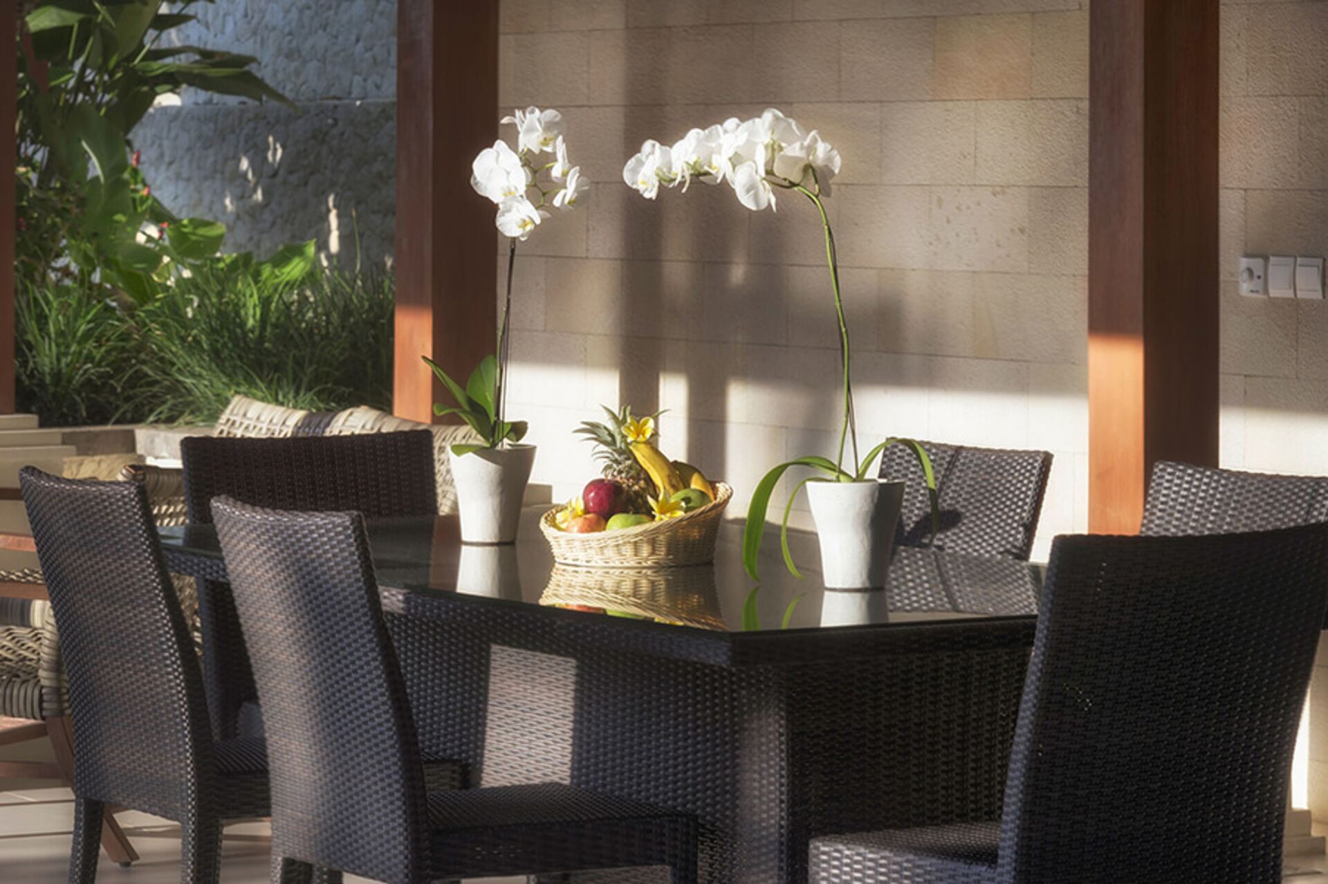 A dining area with wicker chairs and a table, a potted orchid, and a fruit basket.