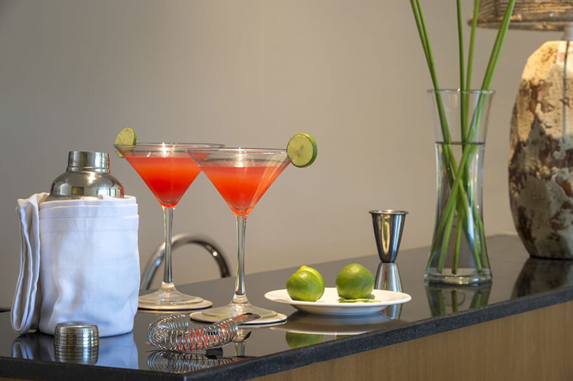 Eine Bar mit zwei Martini-Gläsern, einem Cocktail-Shaker, einer Spirituosenflasche, einem Teller mit zwei Limetten und einer Vase mit grünen Stielen.