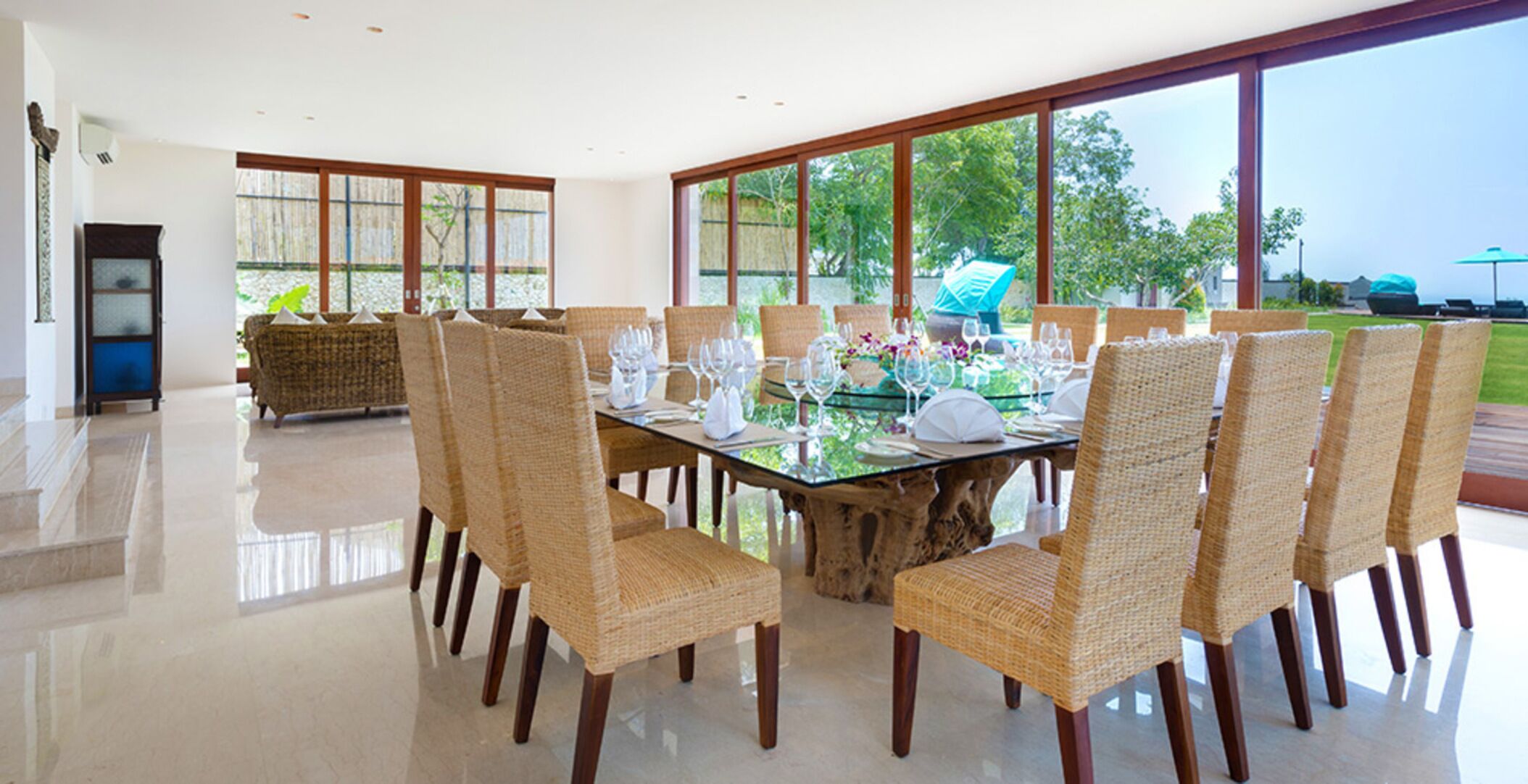A dining area with a glass table surrounded by wicker chairs, large windows, and a view of greenery outside.