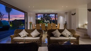 Una sala moderna con muebles de mimbre, un comedor con vista al atardecer y una escalera que conduce a un nivel superior.