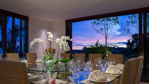 Mesa para seis personas dispuesta con vajilla blanca, rodeada de sillas de mimbre, con vistas a un paisaje montañoso al atardecer.