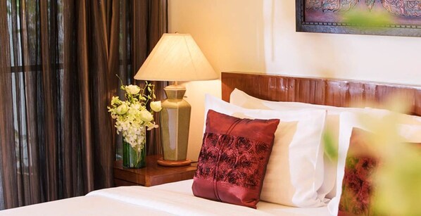 A neatly made bed with white linens and a red patterned pillow, a green vase with white flowers, and a table lamp on a wooden nightstand.