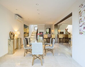 A modern dining area with a glass table, wooden chairs, and a marble floor. There is a staircase in the background leading to another level.