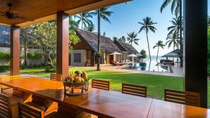 Una mesa de comedor de madera con sillas y una cesta de frutas, con vistas a una zona de piscina con palmeras y un edificio con techo de paja.