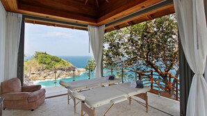 Une pièce baignée de lumière naturelle, avec un plafond en bois, une table de massage, un fauteuil confortable et une vue sur un paysage côtier.