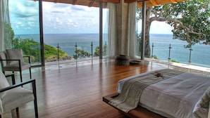 Une chambre avec une grande fenêtre donnant sur la mer, un lit, une chaise et un petit coin salon.