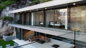 Een modern huis met een glazen balkon, een zithoek met een bank en salontafel, en een slaapkamer met een bed en nachtkastje.