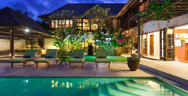 A poolside area with lounge chairs and a swimming pool, surrounded by lush greenery and a building with large windows.