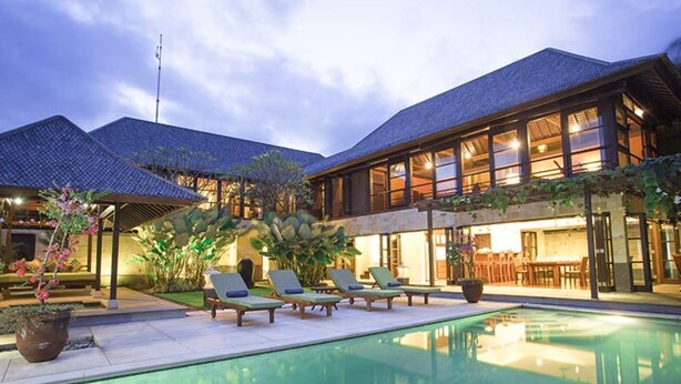 A poolside area with lounge chairs and a swimming pool, surrounded by a building with large windows and a tiled roof.