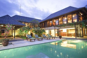 Zona de piscina con sillones de descanso y una pileta, rodeada por un edificio con grandes ventanales y un techo de tejas.
