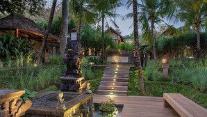 A wooden deck leading to a traditional house with a thatched roof, surrounded by lush greenery and a stone water feature. - Rent a Luxury Villa in Bali Close to the Beach, Bali Villa 1130 (Bali)