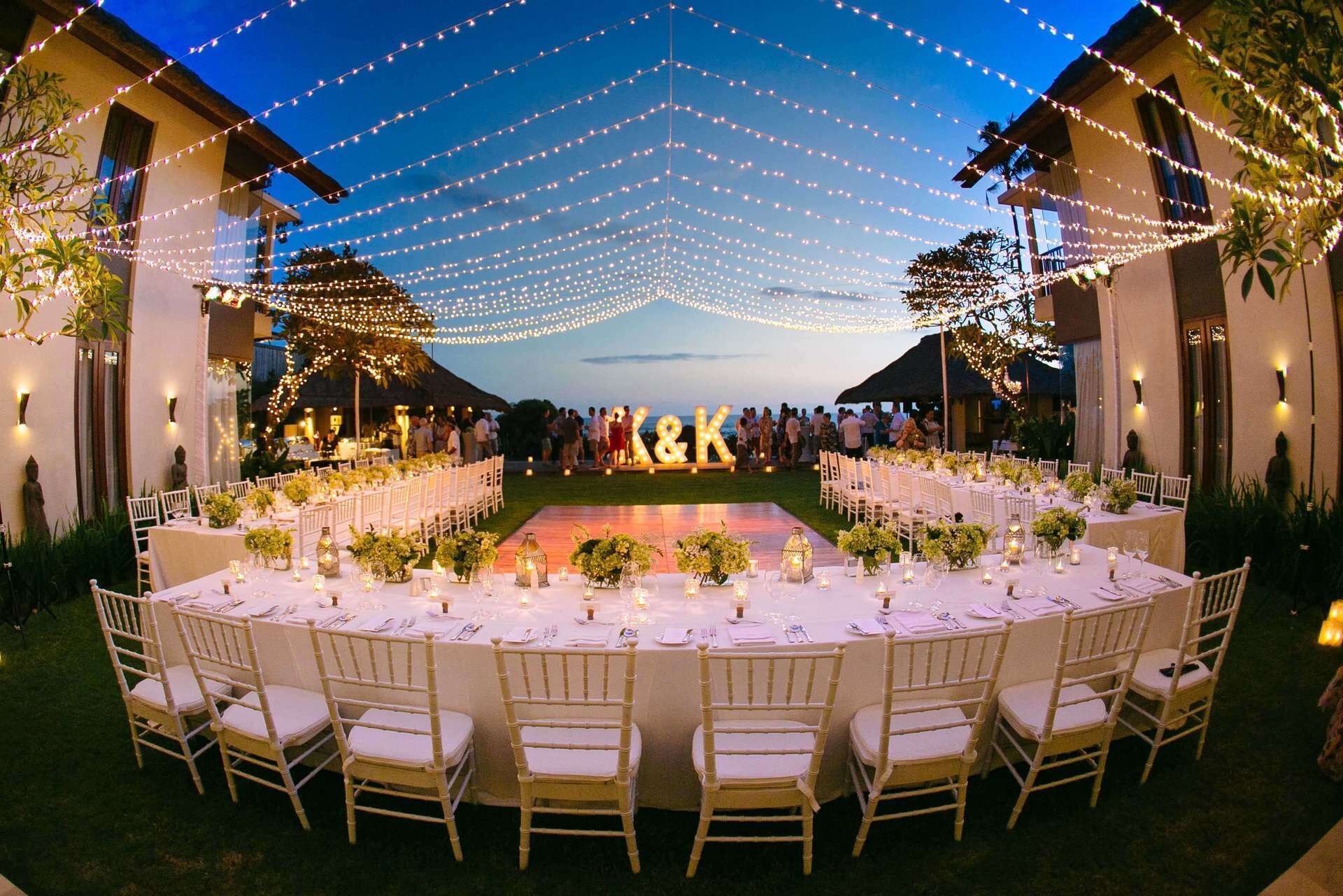 An outdoor wedding reception with round tables, white chairs, and string lights.