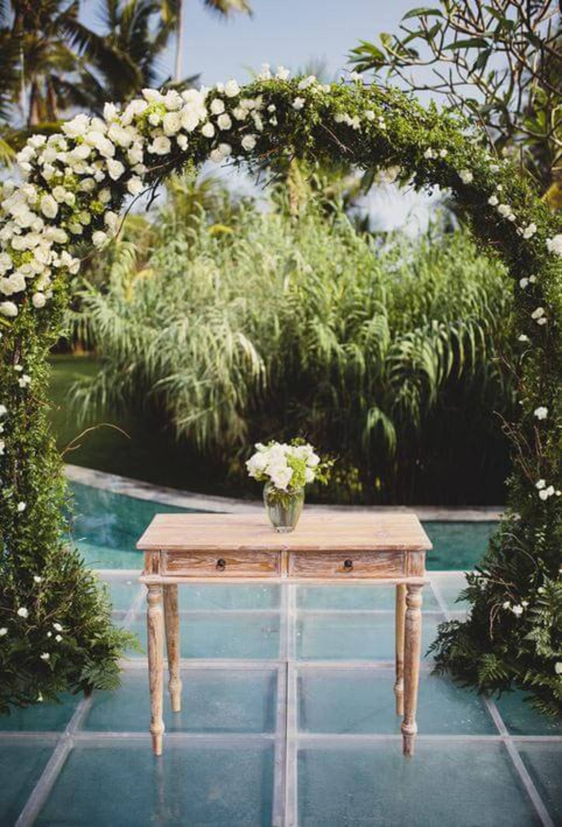 Une table en bois sur laquelle est posé un vase de fleurs, le tout se détachant sur un fond de verdure luxuriante et d’une piscine à l’eau d’un bleu limpide.