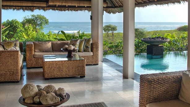 A spacious lounge with wicker furniture, a central coffee table with decorative stones, and a view of the ocean through large windows. - Rent a Luxury Villa in Bali Close to the Beach, Bali Villa 1130 (Bali)