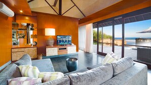 A modern living room with a large sofa, a coffee table, and a TV. There is a view of the ocean through large windows.