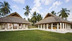 Two thatched-roof structures with white pillars, a lawn, and palm trees in the background.