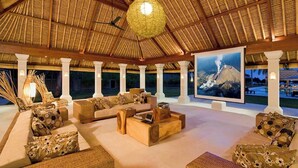 A spacious outdoor lounge area with comfortable seating, a large painting of a mountain, and a thatched roof structure.
