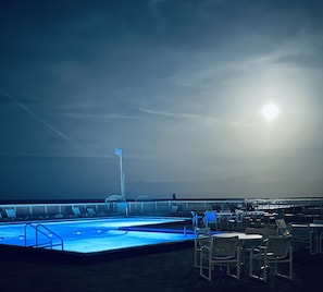 Zona de piscina con sillones de descanso y mesas, un asta de bandera y vista al mar bajo un sol radiante.