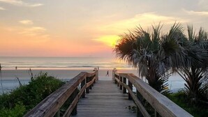 Ein hölzerner Steg führt zu einem Strand, im Hintergrund ein Sonnenuntergang.