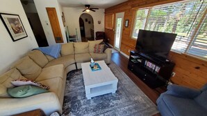 A cozy living room with a sectional sofa, a coffee table, a TV stand with a flat-screen TV, and a large window with blinds. - Country Home at the Crossroads (Springville)