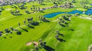 Vista aérea de un campo de golf con varios hoyos, obstáculos de agua y una zona residencial al fondo.
