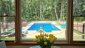 A view of a swimming pool with a net fence, surrounded by trees and a grassy area with chairs. - Modern Cape, Private Heated Salt Water Pool,  Minutes to Beaches/Golf/Lakes (Brewster)