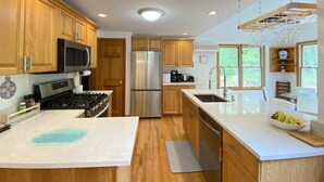 A modern kitchen with wooden cabinets, stainless steel appliances, and a large island with a blue mat. - Modern Cape, Private Heated Salt Water Pool,  Minutes to Beaches/Golf/Lakes (Brewster)