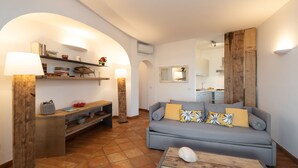 A living room with a grey sofa, wooden side tables, a coffee table with a shell, and a kitchen in the background.