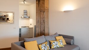 A modern living room with a grey sofa, yellow and black floral cushions, a wooden accent wall, and a kitchen in the background.