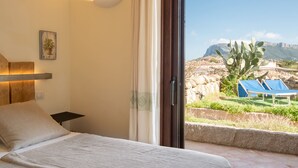 Une chambre à coucher avec un lit, une table de chevet, une lampe fixée au mur et une vue sur un jardin agrémenté d’un cactus et d’une chaise bleue.