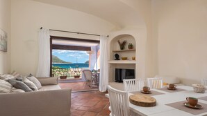 Una sala de estar con un sofá, una mesa de comedor con sillas, un televisor y una chimenea. Se ve el mar a través de un ventanal grande.