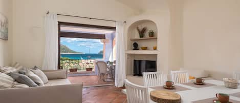 A living room with a sofa, a dining table with chairs, a TV, and a fireplace. There is a view of the sea through a large window.
