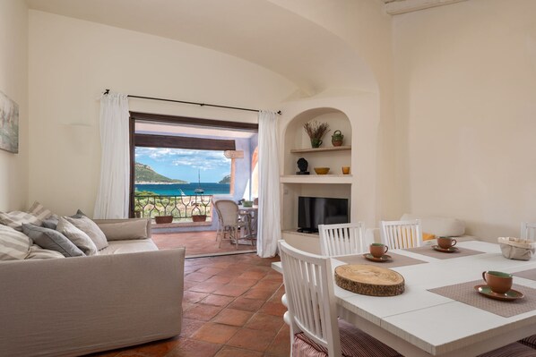 A living room with a sofa, a dining table with chairs, a TV, and a fireplace. There is a view of the sea through a large window.