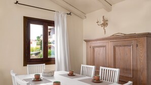 Un comedor con una mesa blanca dispuesta para seis personas, rodeada de sillas blancas. A la derecha, un mueble de madera, y se ve una ventana con cortina.