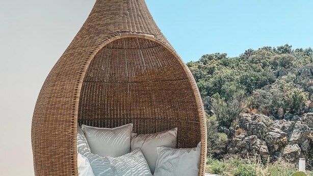 A wicker hanging chair with cushions, a potted cactus, and a rocky hillside in the background.