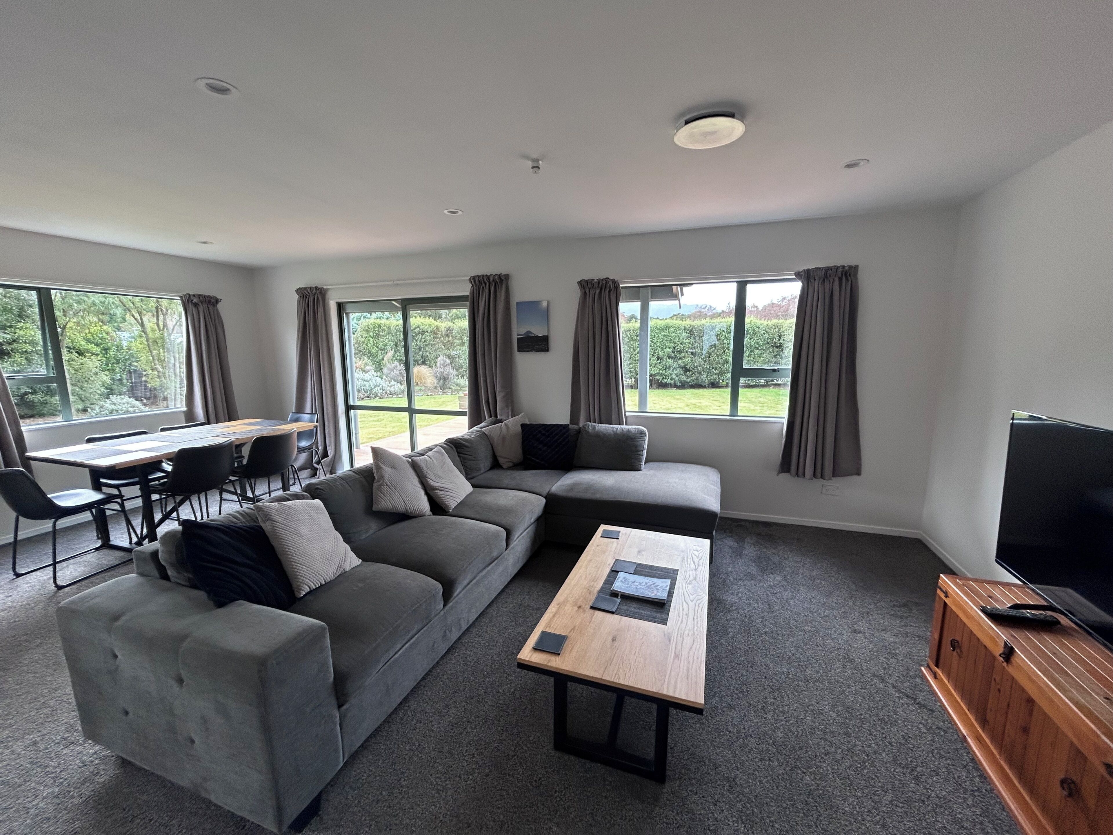 A modern living room with a sectional sofa, a coffee table, a TV, and large windows offering a view of the outdoors.