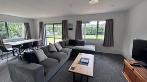 A modern living room with a sectional sofa, a coffee table, a TV, and large windows offering a view of the outdoors. - Alpine Bach with spa and separate games room. (Ohakune)
