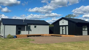 Two small, single-story buildings with flat roofs, a grassy area in front, and a clear blue sky with clouds. - Alpine Bach with spa and separate games room. (Ohakune)