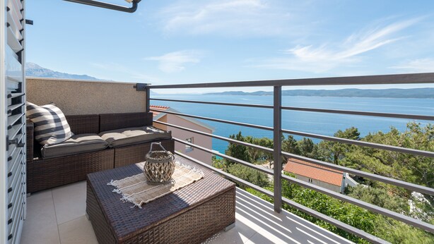 A balcony with a wicker sofa, a small table, and a view of the sea and mountains. - Two bedroom apartment with wonderful sea view (Mimice)