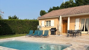 Una piscina con sillas de descanso, una zona de barbacoa y una mesa de comedor al aire libre, frente a una casa con techo de tejas.
